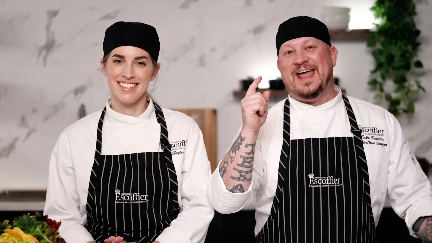 Two smiling Escoffier chef instructors hosting a Studio E live session at a kitchen counter with a laptop.