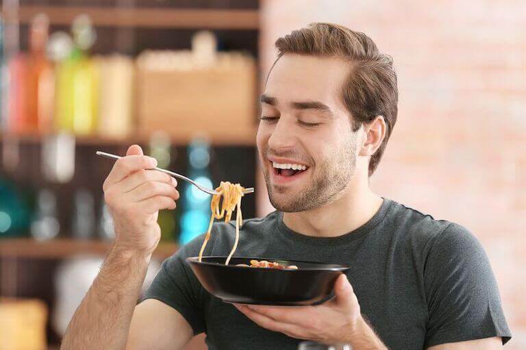A man smiling and eating pasta from a black bowl
