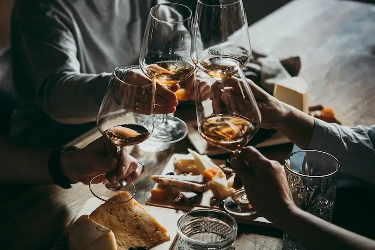 Four people raising glasses of white wine above a table set with cheese, bread, and small bites.