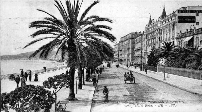 An image of a 19-century black-and-white postcard depicting a street lined on one side by buildings with an ornate, French architectural style, and on the other by palm trees and a seaside promenade with many pedestrians.
