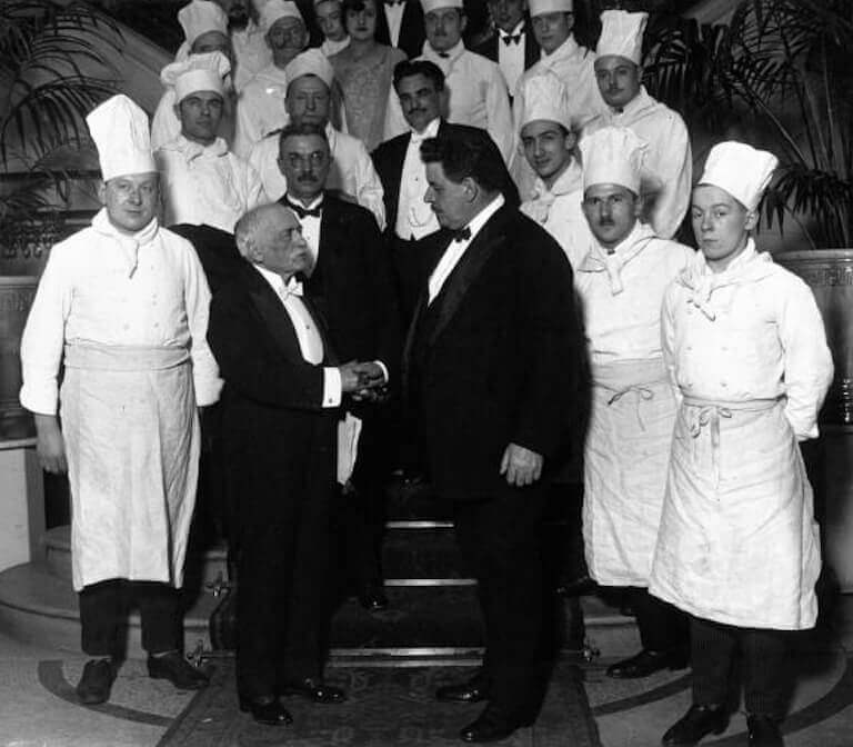 Escoffier and a taller man, both wearing tuxedos, shake hands while surrounded by several other people, some wearing tuxedos and others wearing white chef's uniforms.