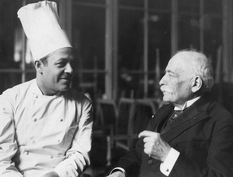 Escoffier, dressed in a dark suit, engages in a friendly discussion with a chef wearing a white uniform and tall chef’s toque while seated in a restaurant dining room.