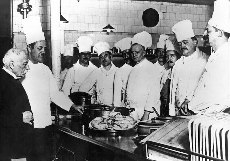 Standing in a professional kitchen, Escoffier looks on as a chef de cuisine displays a platter of whole roast poultry, while ten other chefs in white uniforms stand by and watch.