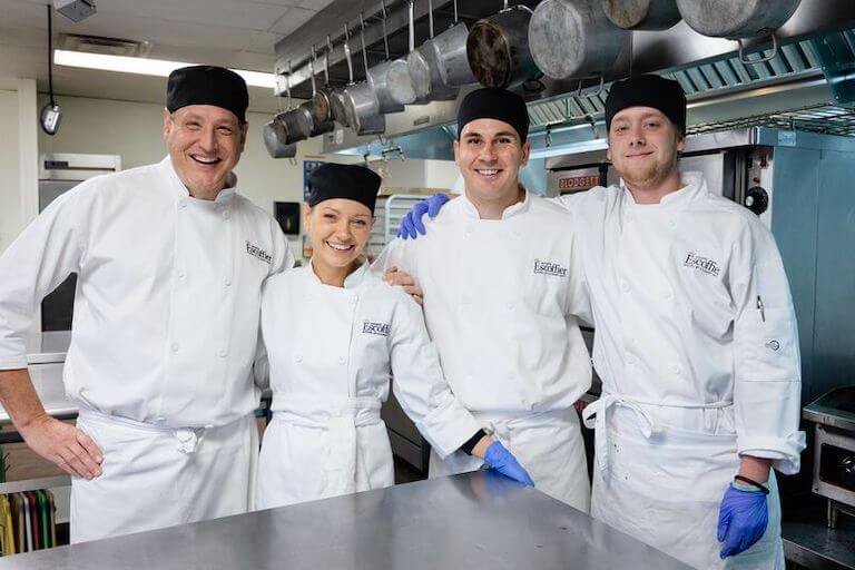 A group of four culinary school students wearing white chef uniforms in a training kitchen.