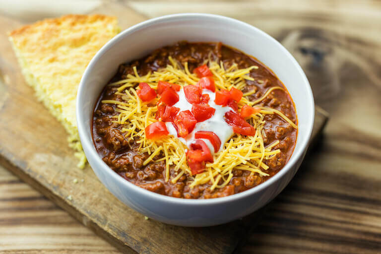 An example of chili con carne showing a bowl of rich, deep-red stew topped with grated cheddar cheese, a dollop of sour cream, and diced tomatoes.