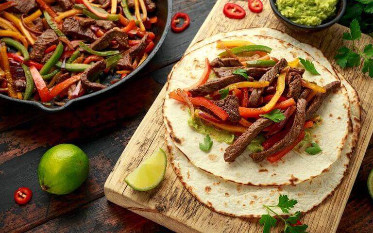 An example of fajitas, showing bright yellow, green, and red bell peppers and grilled beef strips, served on a lightly browned flour tortilla and a wedge of lime.