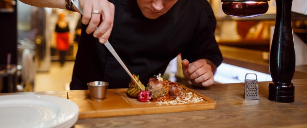 Chef using metal tool to plate dish on a wooden tray 