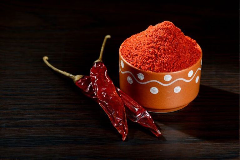 Two dried red chile peppers next to a small earthenware bowl overfilled with red chili powder.
