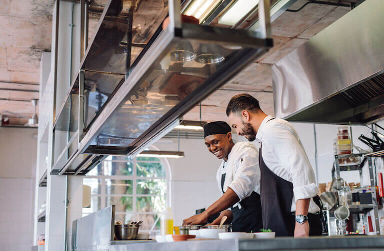 Two male chefs smiling and laughing as they work together in a restaurant kitchen.