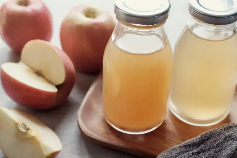 Apple cider vinegar with mother in glass bottles on a wooden cutting board next to sliced apples.