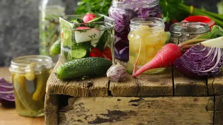 Jars of pickled vegetables, including cucumbers, radishes, cabbage, and garlic, displayed on a rustic wooden surface with fresh produce nearby.