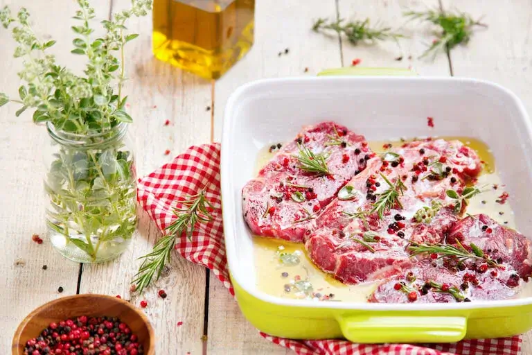 Raw steaks marinating in a dish with vinegar, herbs, and peppercorns, surrounded by fresh rosemary, oregano, olive oil, and a red checkered cloth.