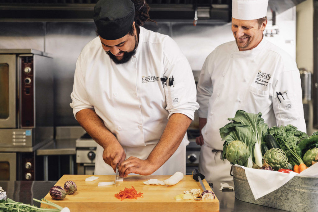 Male culinary student cutting vegetables while smiling chef instructor observes