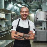 young chef in apron keeping tattooed arms crossed and smiling while standing in a restaurant kitchen