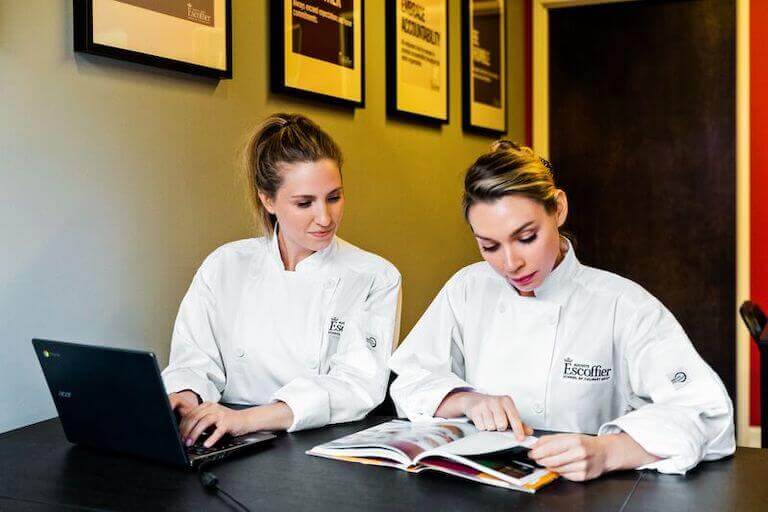 Two Escoffier students studying, one is typing on a computer as the other reads from a book