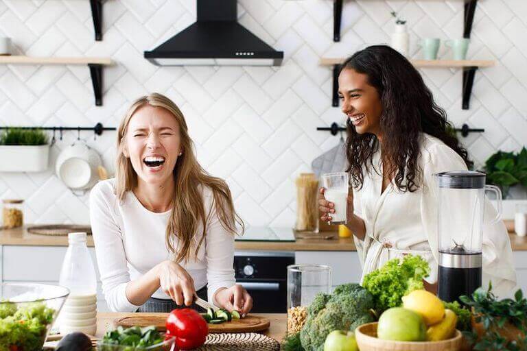 Two woman laughing in the kitchen while one cuts a cucumber and the other is holding a glass of milk