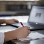 student taking notes on notepad with red pen while looking at computer