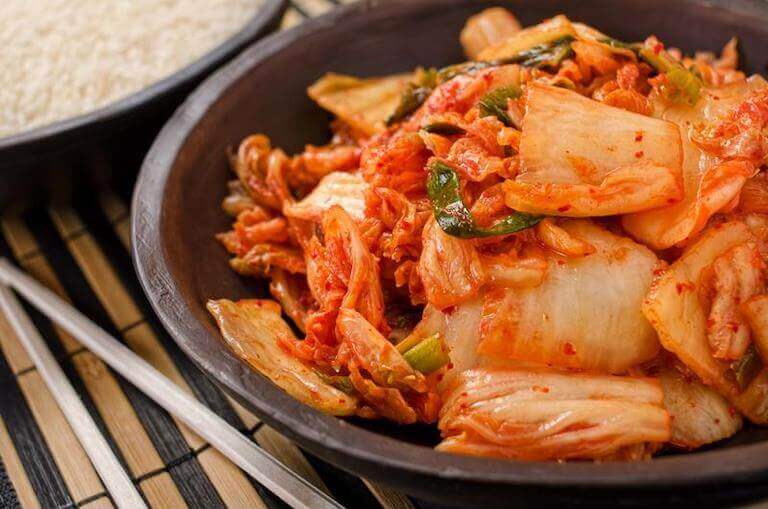 A black bowl of kimchi—fermented cabbage and other vegetables bathed in chili sauce—sits next to a pair of chopsticks and a bowl of rice.