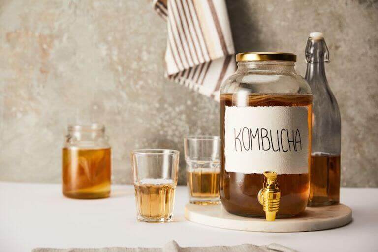 A large jug labeled “Kombucha” sits on a countertop next to some bottles and glasses all containing the same amber liquid.