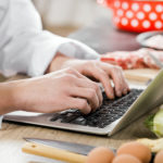 Chef typing on laptop computer on table with knife, eggs, and vegetables