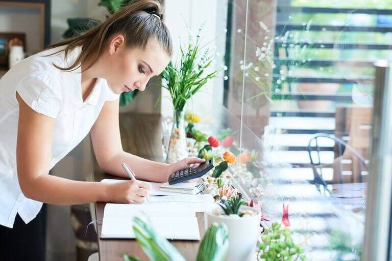 A woman in a white shirt stands by a window and uses a calculator to check business accounts.