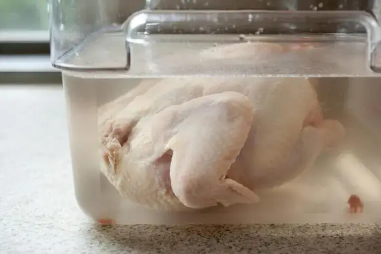 A whole raw chicken submerged in a clear container of brine solution on a kitchen countertop.