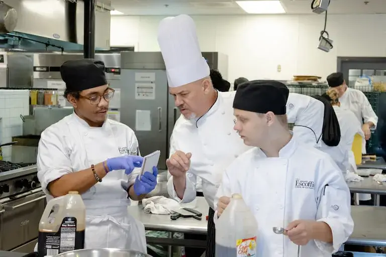 An Escoffier chef instructor wearing a tall white toque reviews a student's handwritten notes with them and another classmate at a stainless steel kitchen prep table.