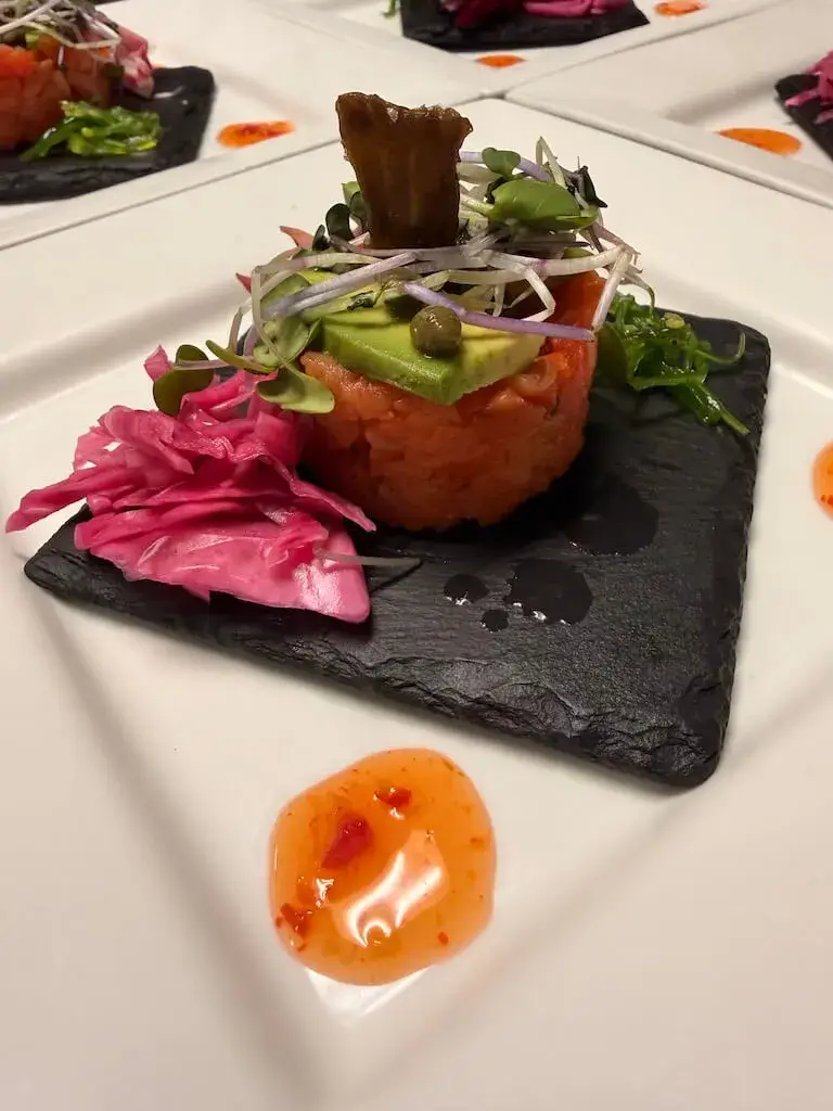 An elegantly plated salmon tartare tower topped with sliced avocado and microgreens. It is served on a small black slate tile with pickled pink cabbage, seaweed salad, and a side of sweet chili sauce.