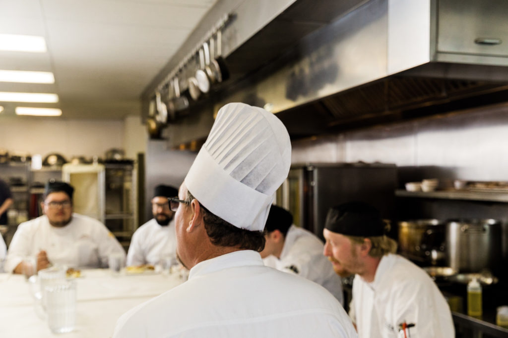 Chef talking to group of male cooks and culinary students in a kitchen