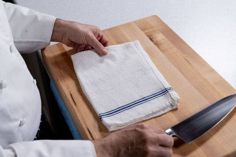 A pair of hands, the right one holding a chef’s knife and the left hand holding a dish towel resting on a large wooden cutting board.