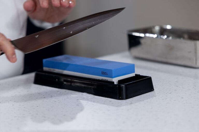Closeup of a pair of hands holding a chef’s knife at an angle over a blue whetstone.