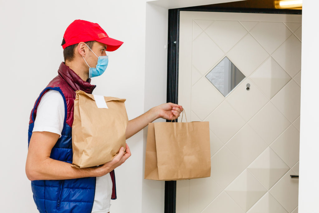 Delivery man with red hat and mask holding paper bag with food on white background