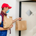 Delivery man with red hat and mask holding paper bag with food on white background