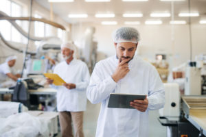 Man in white lab coat and hairnet standing and holding tablet