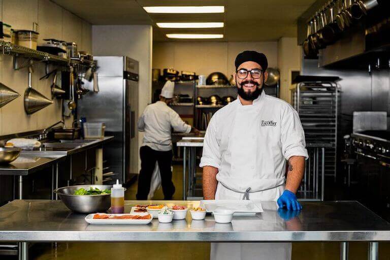 Escoffier student smiling for a picture as they stand behind a metal table with salad ingredients