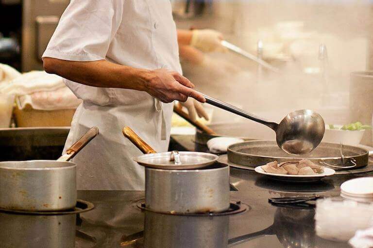 A chef stirs a pot with a large ladle in a busy restaurant kitchen filled with steam.