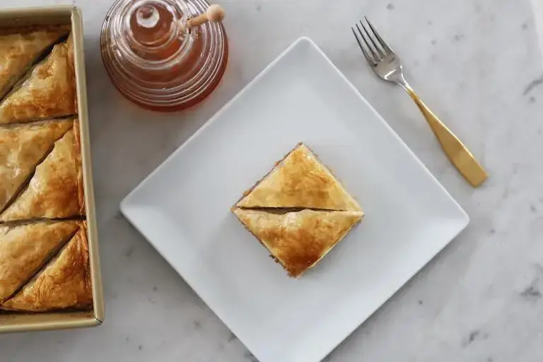 An overhead view of a piece of baklava on a square white plate with a jar of honey, a fork, and a pan filled with more baklava.
