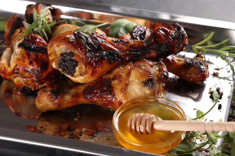 Close-up of glazed roasted chicken drumsticks on a metal tray, garnished with herbs and served alongside a small glass bowl of honey with a honey dipper.