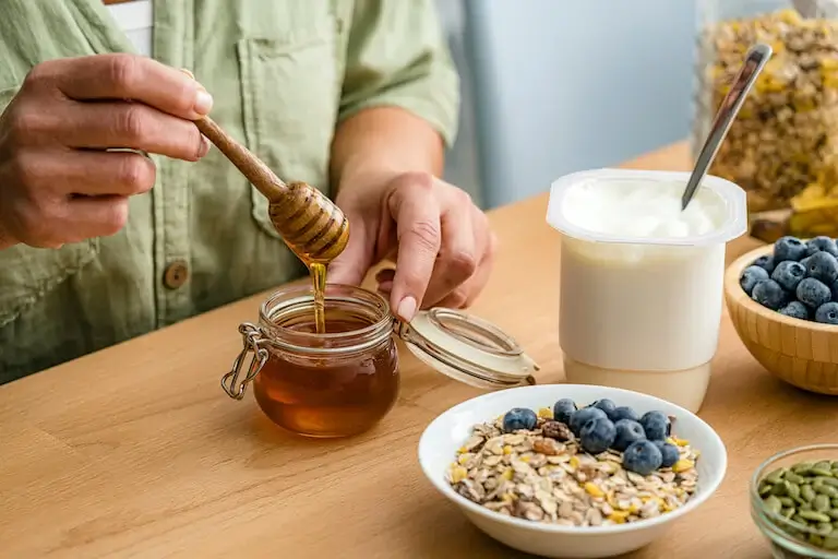 Person drizzling honey from a dipper into a glass jar, with yogurt, blueberries, and a bowl of granola and seeds arranged on a wooden table.