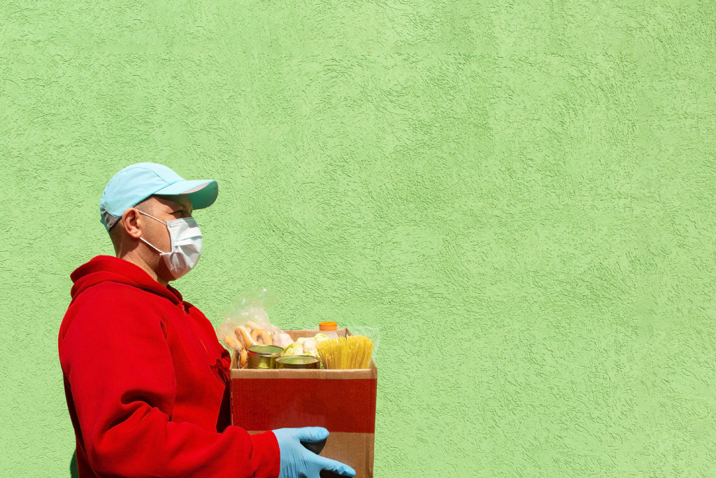 man with red hoodie, mask, and blue hat holding a box of food, help and donation of food 