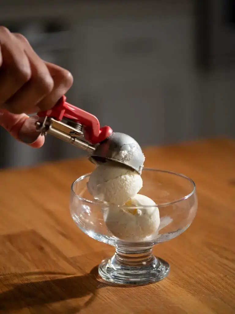 Hand scooping vanilla ice cream into a glass dessert dish with a metal scoop.