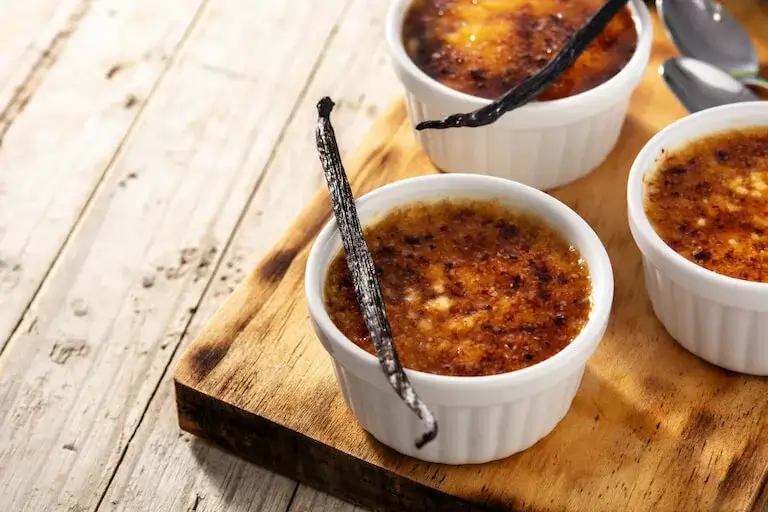 Three ramekins of crème brûlée topped with caramelized sugar and garnished with whole vanilla beans, sitting on a wooden board.