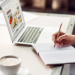 Student taking notes of food in journal while using laptop computer