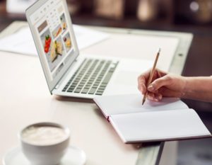 Student taking notes of food in journal while using laptop computer