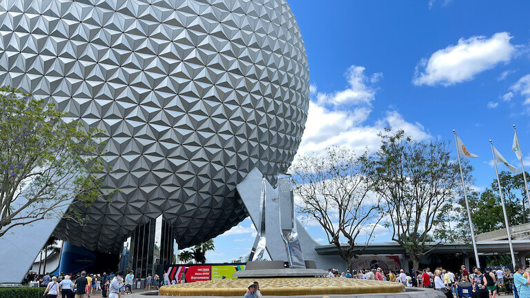 A crowd gathers in the shadow of Epcot, its large spherical shape looming on the left side of the image.