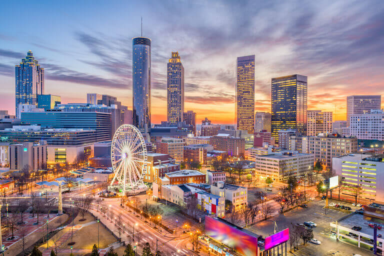 The Atlanta skyline set against a sunset with buildings and a ferris wheel lit up for the evening.