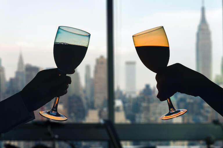 The silhouette of two wine glasses raised in a toast in front of a window through which stands the New York City skyline.
