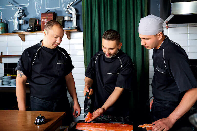 Head chef displaying a filet of raw salmon to two other chefs in a restaurant kitchen.