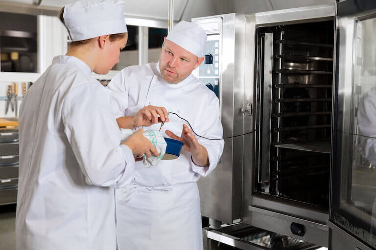 Chef training a new employee while hey hold a casserole dish in front of a large oven