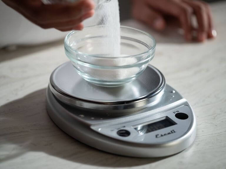 A small kitchen scale sits on a counter as sugar pours into a clear glass bowl resting on its surface.